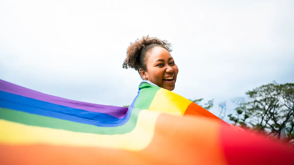 A Black woman smiles at the camera while draped in a rainbow flag.